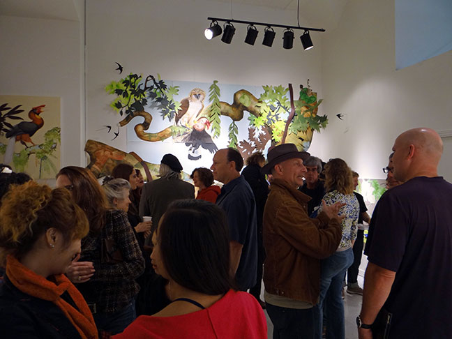 Visitors mingle beneath spotlights in a crowded gallery, with colorful artwork of birds, twisting branches, and foliage displayed on the walls.