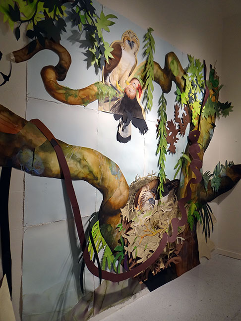David Tomb’s installation depicts a Great Philippine Eagle perched above a chick in a nest, surrounded by twisting branches, hanging vines, and layered cutout foliage extending from the gallery wall.