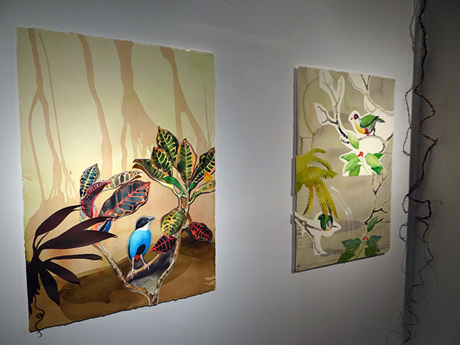 Two paintings by David Tomb hang side by side: a blue-and-black bird among vividly patterned leaves on the left, and two green birds perched among pale branches and foliage on the right.