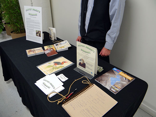 A black-covered table displays Jeepney Projects Worldwide information signs, bird art prints, brochures, and an open handwritten guestbook, with a person standing behind it.