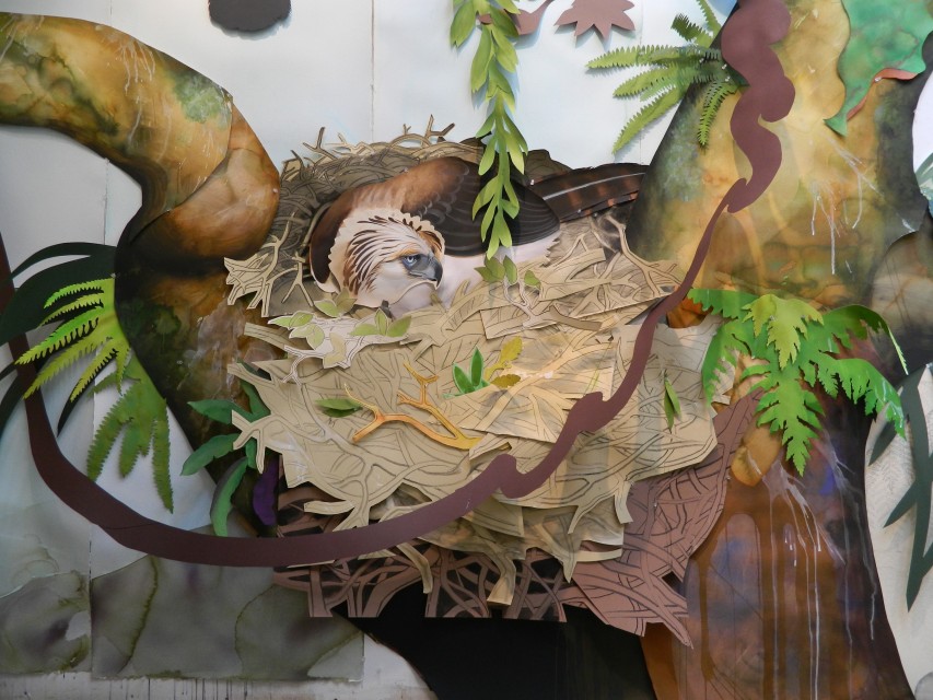 David Tomb installation depicting a Philippine eagle nestled in a large nest of layered cutout branches, surrounded by painted tree trunks, vines, and tropical foliage.